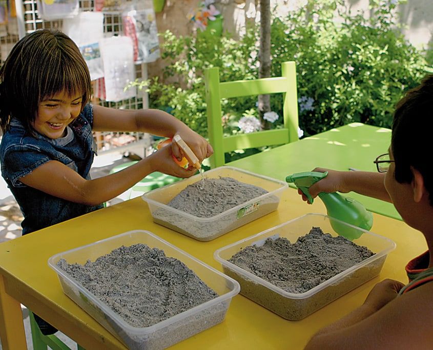 Bahçedeki sarı masada eleklenmiş kumla dolu plastik kaplar, çocuklar kumu nemlendiriyor