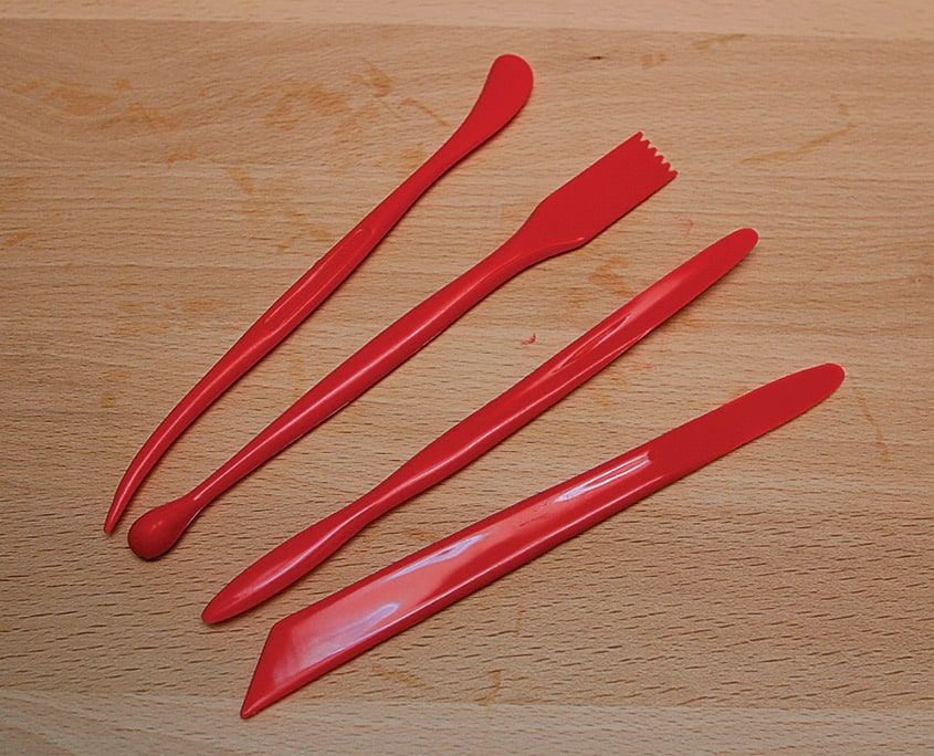 Ahşap zeminde dört kırmızı plastik kil şekillendirme aracı: kaşık, tarak ve spatula uçlu