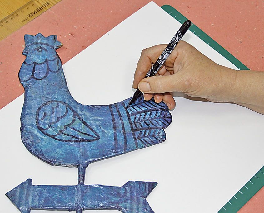 Mavi boyalı horozun kanat ve kuyruk desenleri siyah kalemle çiziliyor, altta ok görünüyor