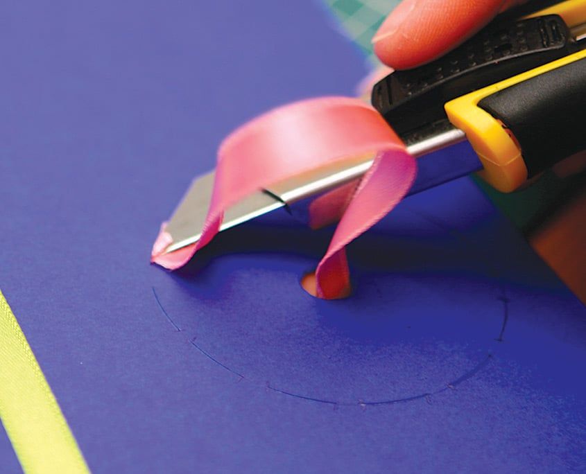 Maket bıçağıyla lacivert kartondaki daire çizgisine yarık açılıyor, pembe kurdele yanında