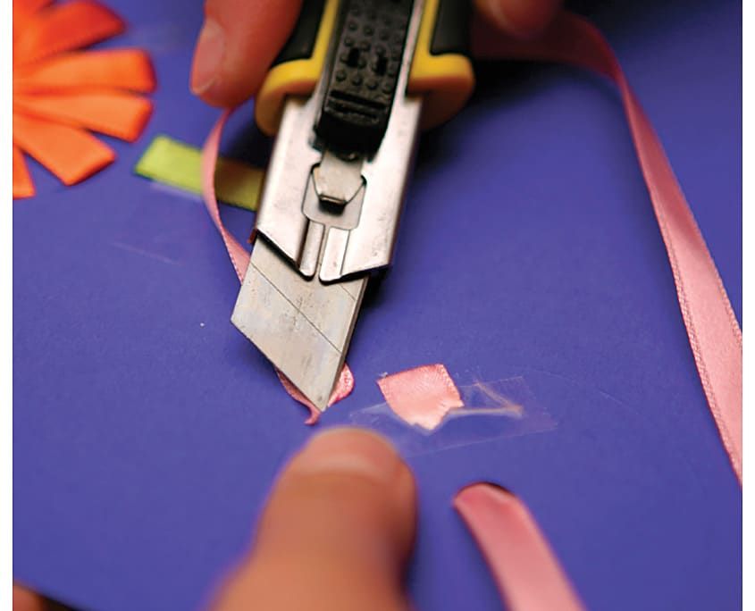 Kartonun arkasında pembe kurdelenin fazlası maket bıçağıyla kesiliyor, uç bantla sabit