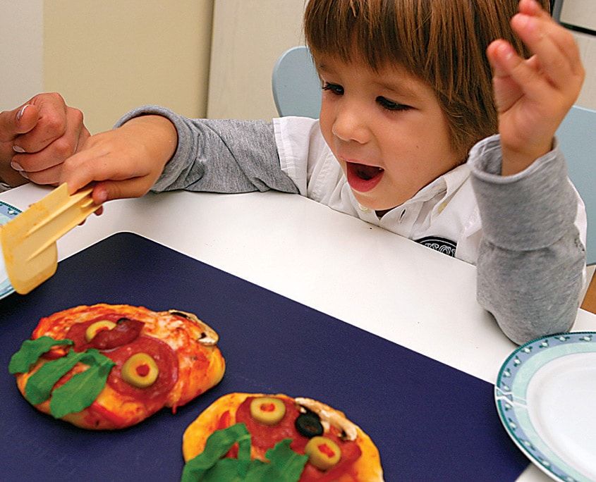 Roka, zeytin ve salamla yüz yapılmış iki pişmiş mini pizza, bir çocuk masadan uzanıyor