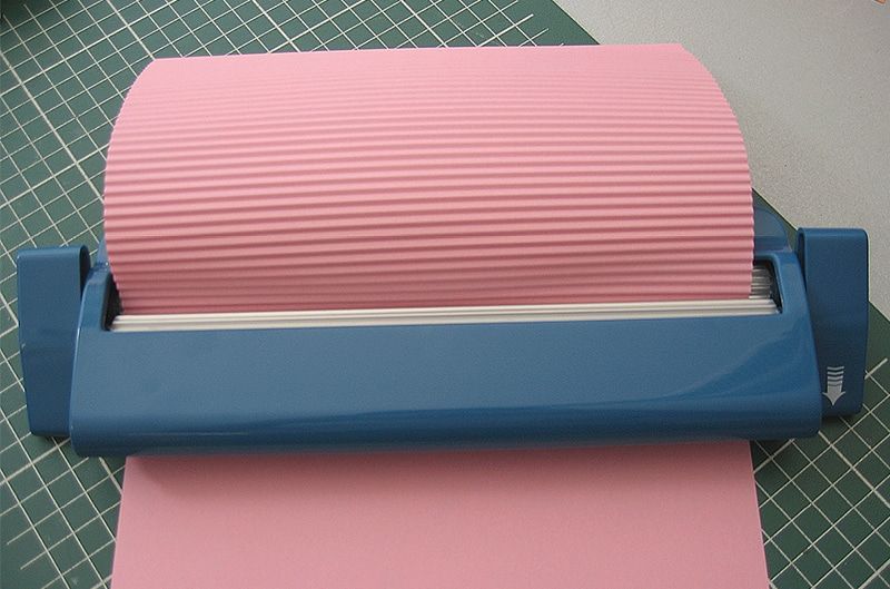 Mavi kağıt kıvırma makinesinden dalgalı olarak çıkan pembe kağıt, kesim matı üzerinde