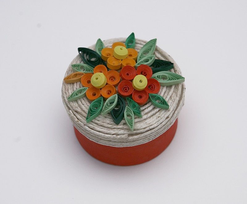 İple sarılmış yuvarlak kutunun kapağında turuncu, sarı ve yeşil quilling çiçek süslemesi