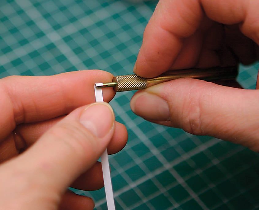 Beyaz quilling şeridinin ucu yarıklı metal dolama aletine takılıyor, arkada yeşil kesim altlığı