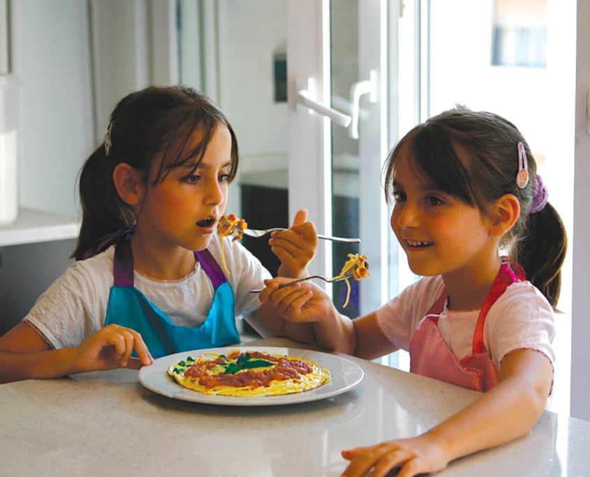 Önlüklü iki çocuk masada tabaktaki makarnalı pancake'i çatalla paylaşarak yiyor