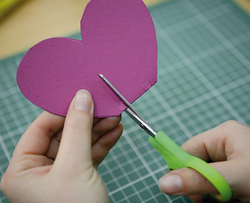 Pembe kartondan kalp biçimli kanadın yeşil saplı çocuk makasıyla kenarından kesilmesi