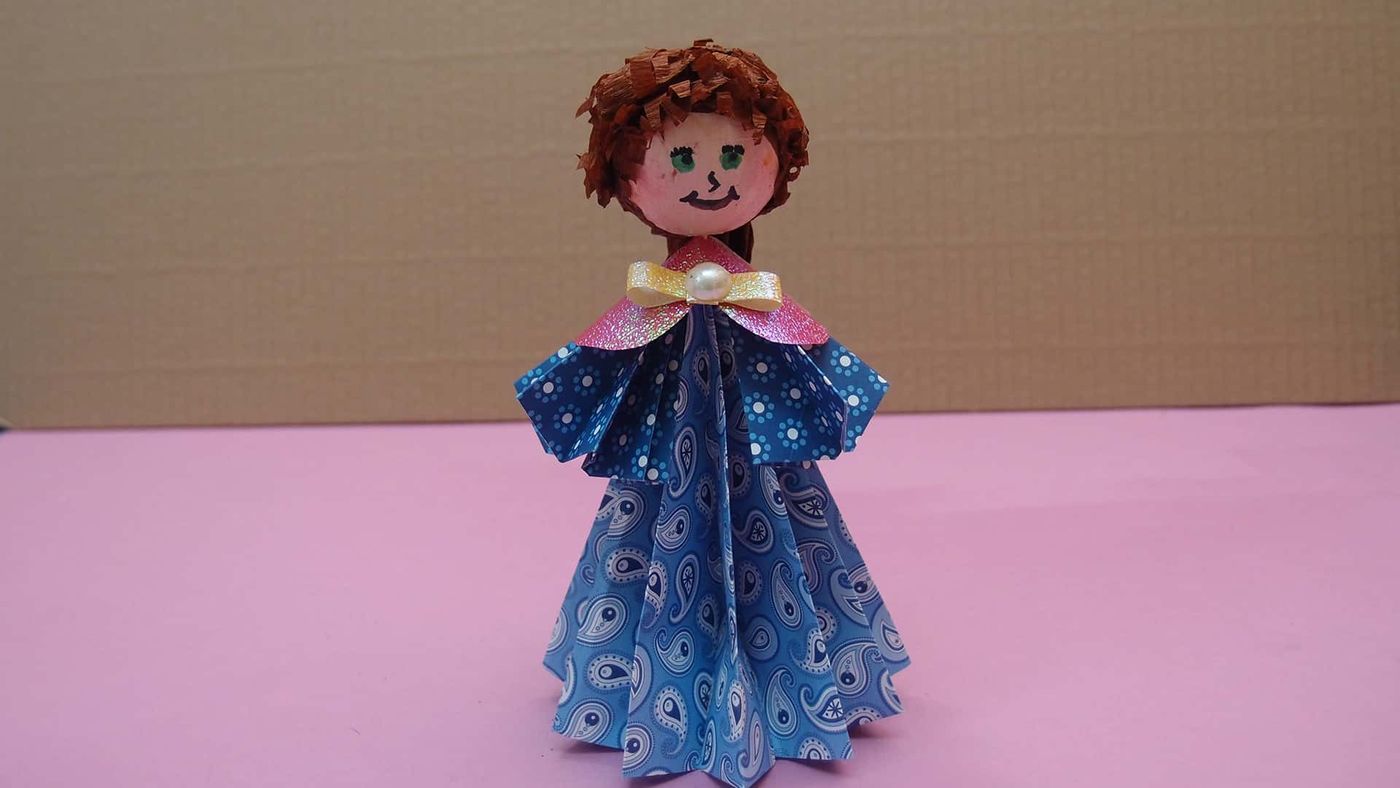 Tamamlanmış origami kız bebek: kahverengi kıvırcık saçlı, pembe yakalı ve mavi desenli elbiseli