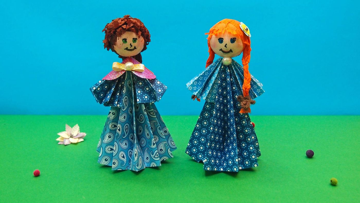 Mavi ve yeşil fon önünde iki origami kız bebek, yerde kağıt çiçek ve renkli küçük toplar