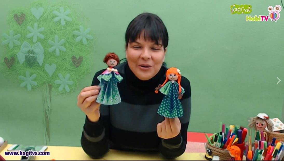 Origami Çocuklar canlı atölye yayınından kare: eğitmen kağıttan yapılmış iki elbiseli kız bebeği gösteriyor