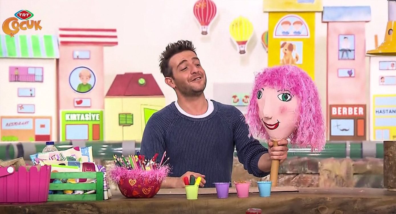 Stüdyo masasında pembe saçlı balon kafa kukla ve tüylü kase kalemlik yan yana, sunucu arkada