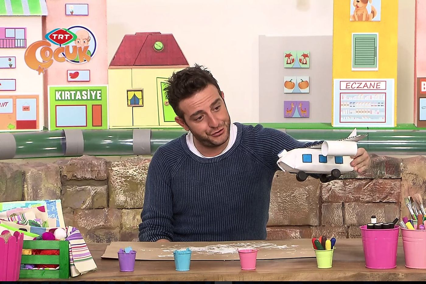 Stüdyo masasında sunucu bitmiş beyaz karton uçağı kaldırıp gösteriyor