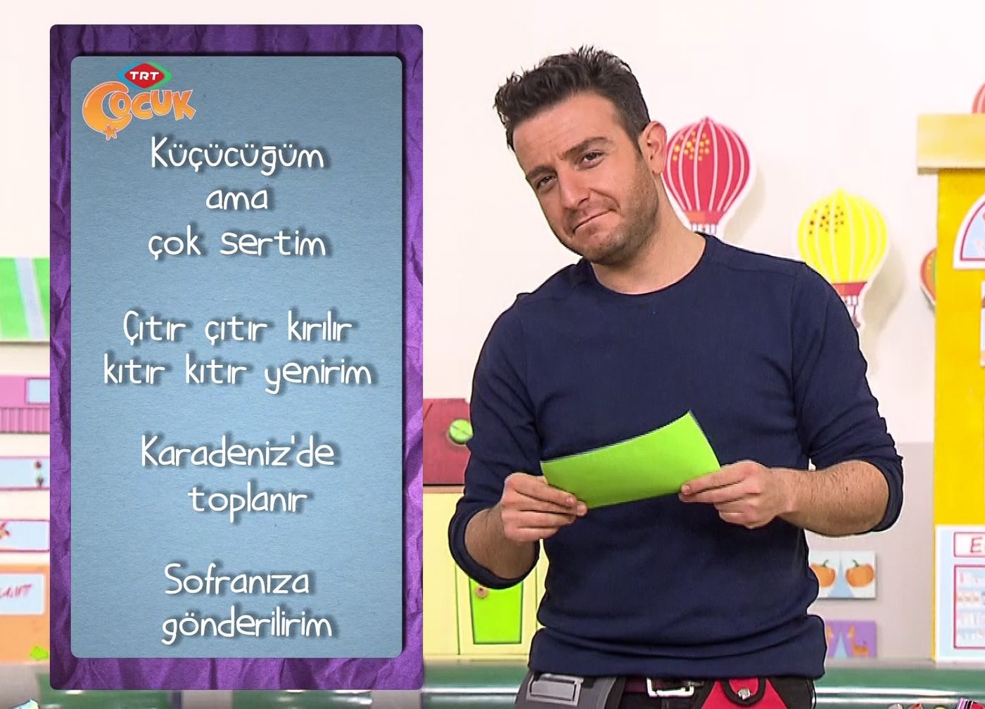 Stüdyoda fındık bilmecesi yazılı mor panonun yanında elinde yeşil kart tutan sunucu