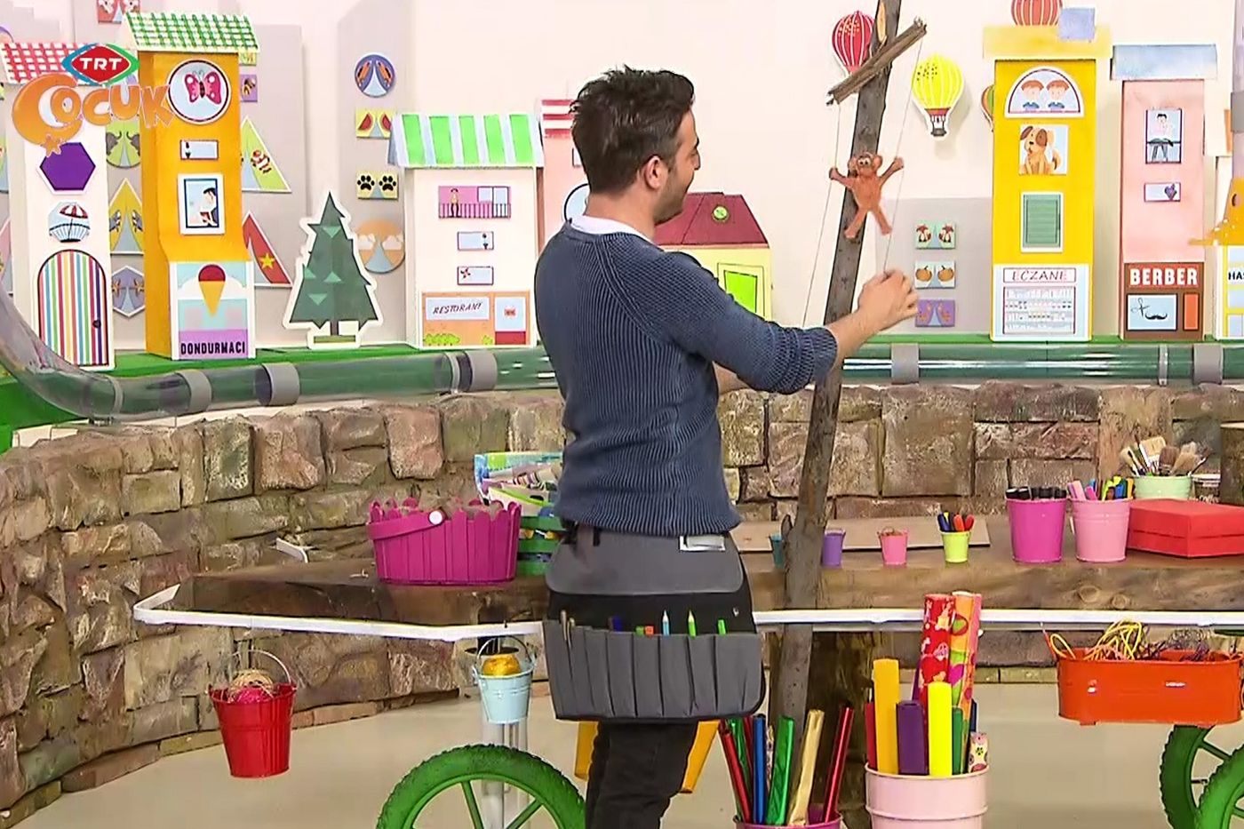 Programdan kare: sunucu stüdyoda dik duran dala bağlı maymunun ipini ayarlıyor