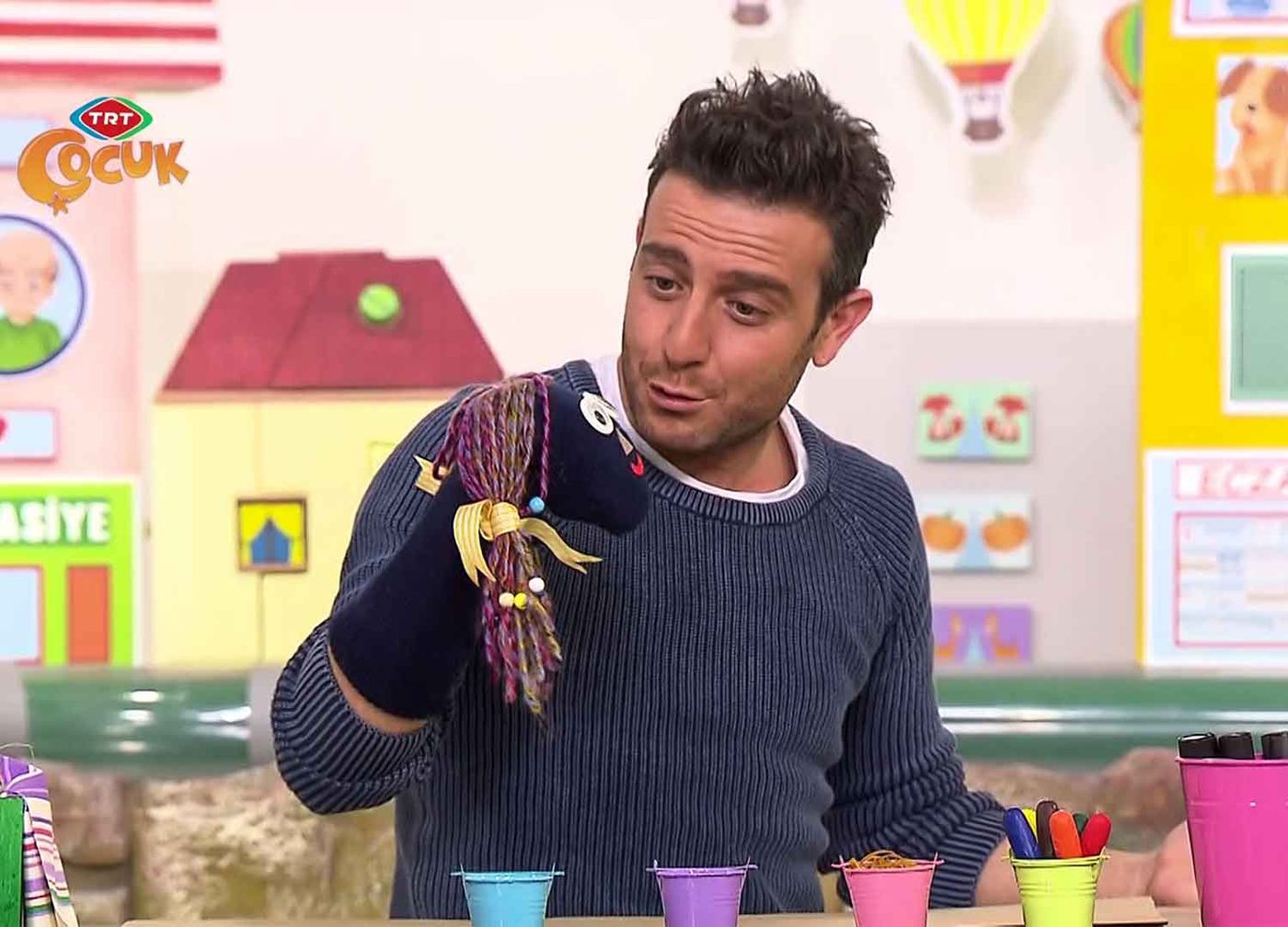 Sunucu eline geçirdiği ip saçlı siyah çorap kuklayı konuşturuyor, önünde renkli kalem kutuları