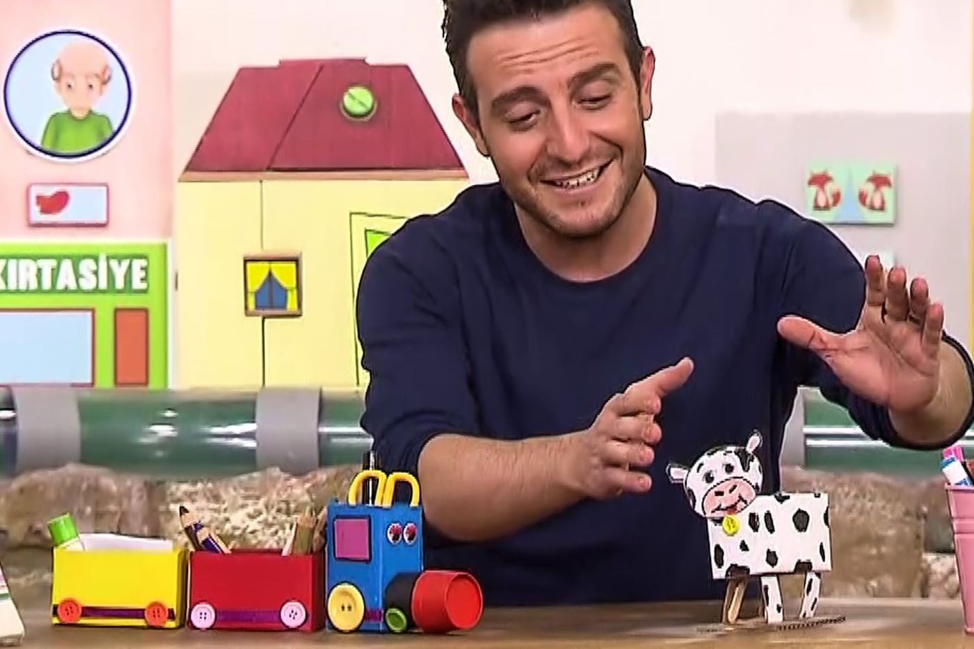 Programdan kare: sunucu kutu tren kalemliği ile benekli ineği masada gösterip anlatıyor