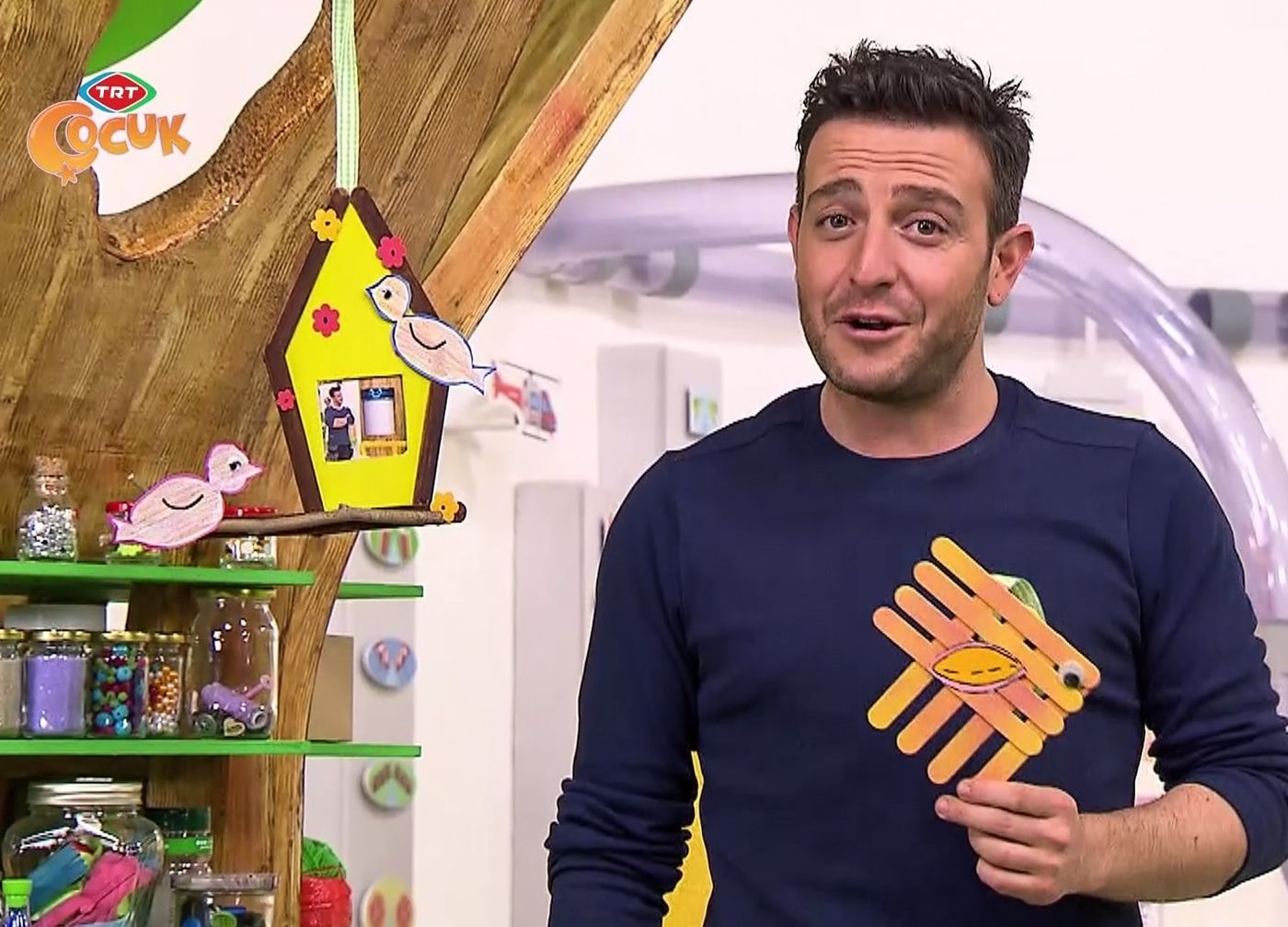Stüdyodan kare: sunucu dondurma çubuklarından boyalı bir parçayı gösteriyor, arkada asılı kuş evi çerçeve