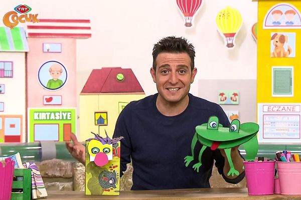 Kitap ayracı yapımı anlatılan programdan kare: sunucu kağıt yüz ayraç ve yeşil kurbağa ayraçla renkli dekor önünde