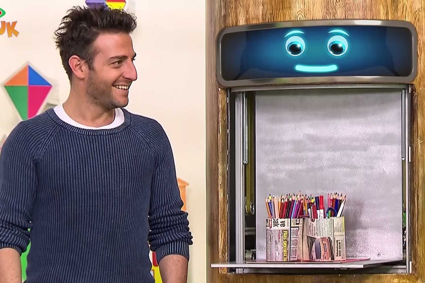 Televizyon programından kare: sunucu stüdyoda, yanındaki pencerede kalem dolu kutular duruyor