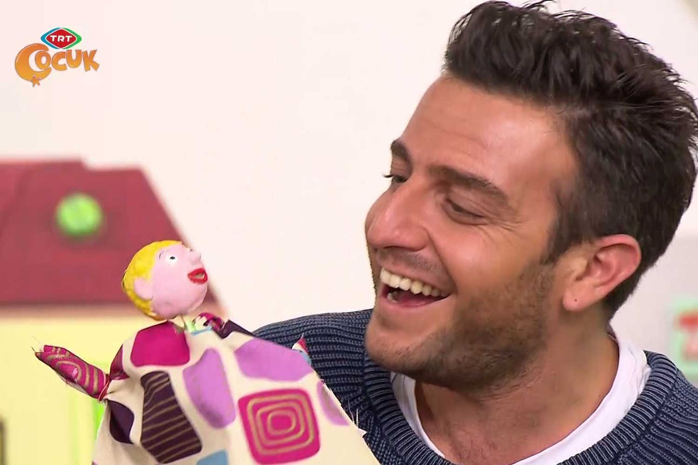 Televizyon programından kare: sunucu gülerek elindeki desenli kumaş kuklaya bakıyor