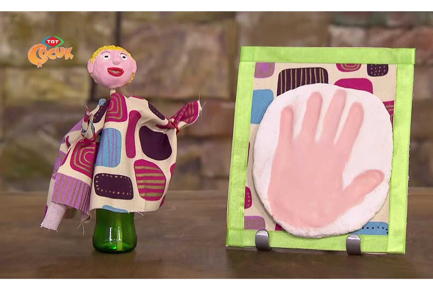 Yeşil şişe üstünde sarı saçlı bitmiş kumaş kukla ve kumaş kenarlı çerçevede pembe hamur el izi
