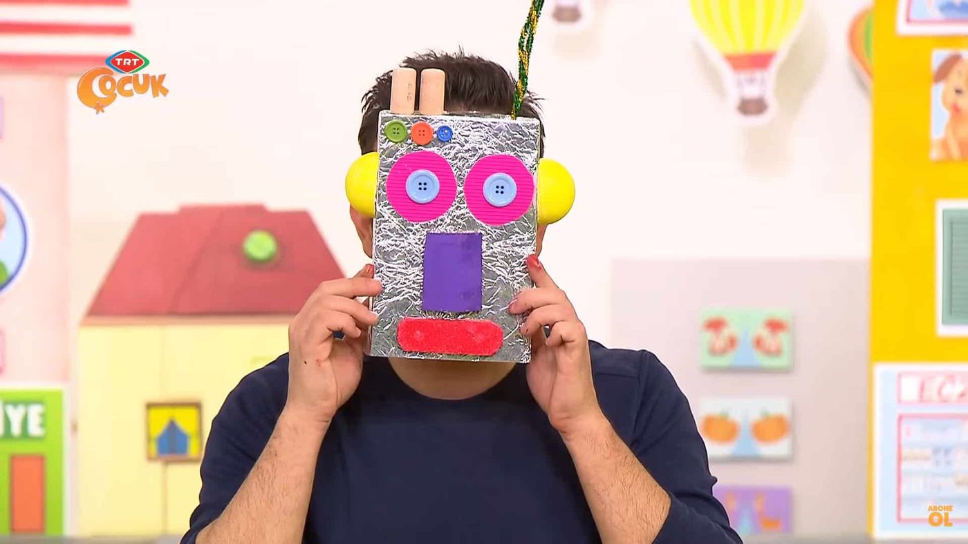 Yayından kare: sunucu pembe gözlü, mor burunlu folyo robot maskeyi yüzünün önünde tutuyor