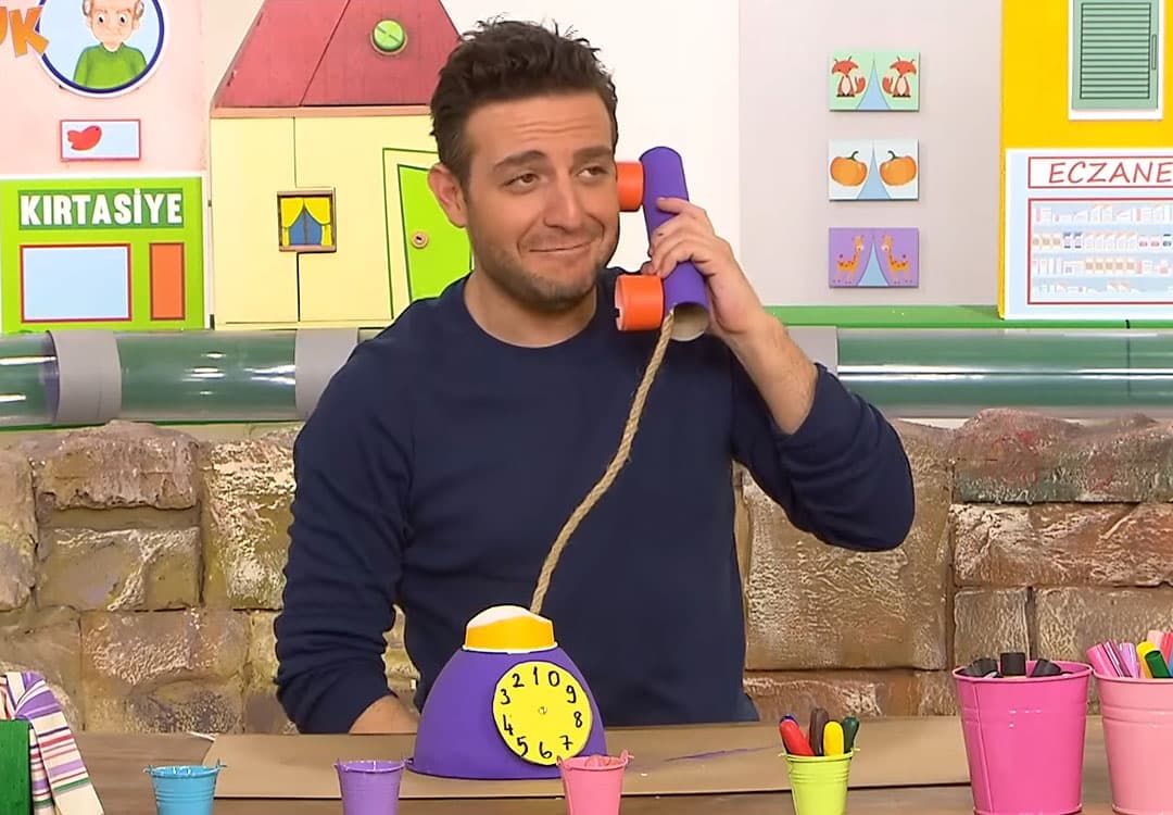 Programdan kare: sunucu kağıt rulo ahizeli oyuncak telefonu kulağına tutuyor, önünde mor telefon