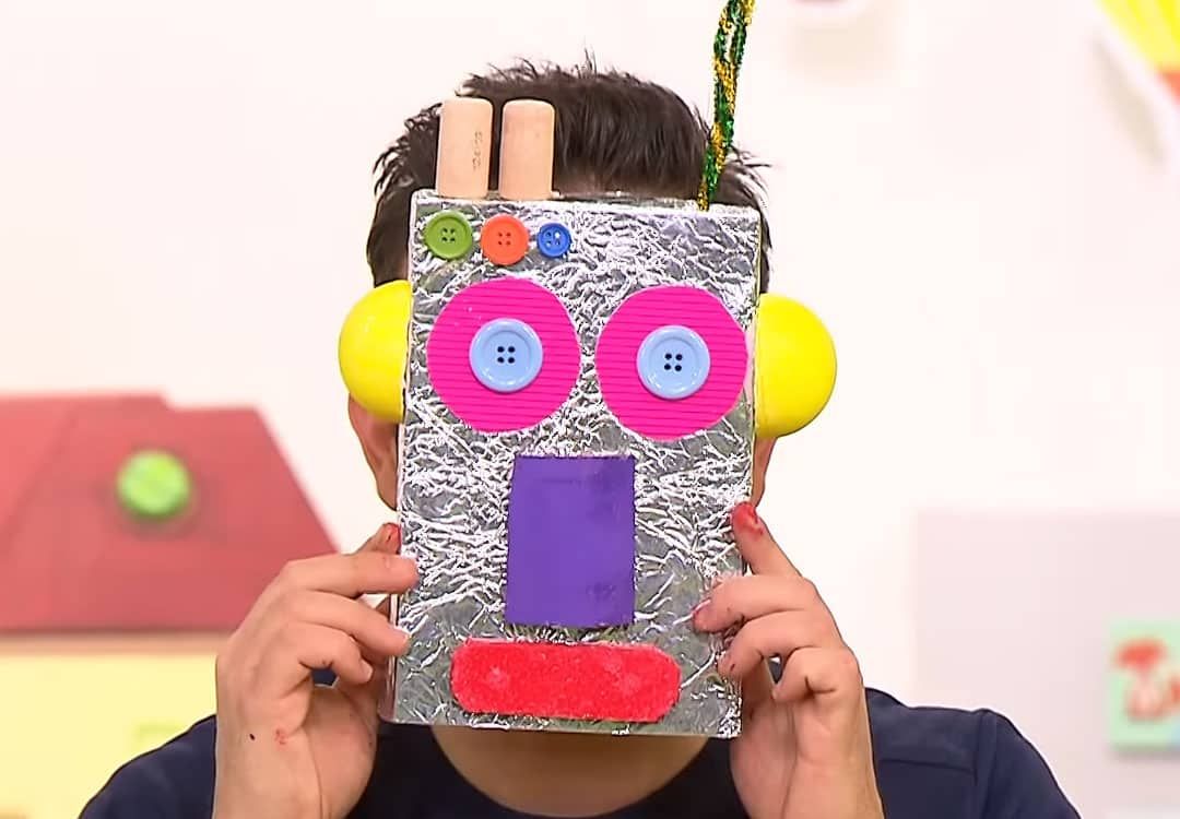 Robot maske yayın karesi: sunucu yüzüne folyo kaplı kutudan, pembe düğme gözlü ve sarı kulaklı maskeyi tutuyor