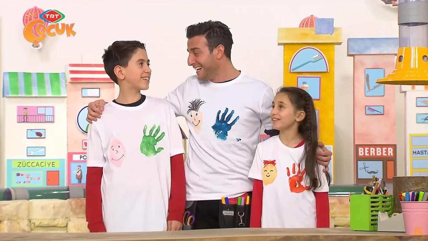 Renkli el izi baskılarıyla süslenmiş beyaz tişörtleri gösteren sunucu ve iki çocuk, stüdyoda yan yana