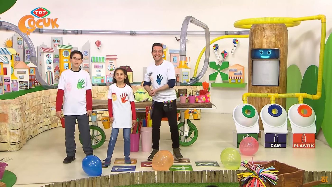 Televizyon stüdyosunda el izi baskılı tişörtleriyle duran sunucu ve iki çocuk, önde renkli balonlar