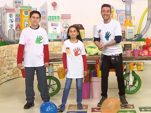 23 Nisan T-shirt boyama: el izi baskılı beyaz tişörtler giymiş sunucu ve iki çocuk renkli stüdyo dekorunda