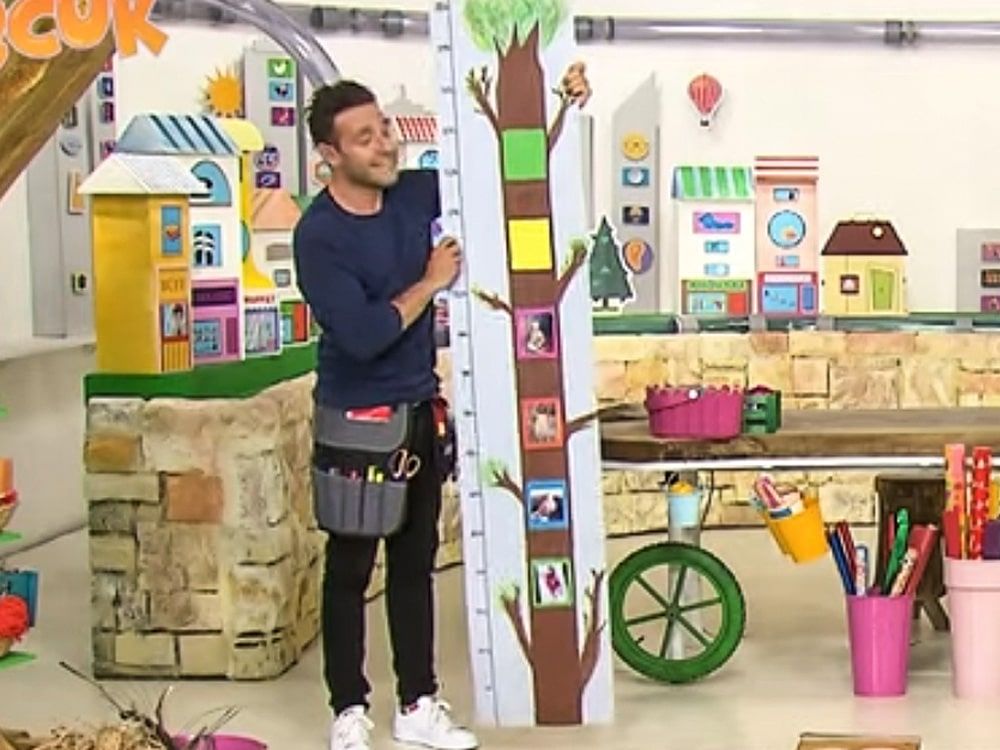 Boy Ölçer: sunucu stüdyoda ağaç gövdesi biçimli, fotoğraf çerçeveli uzun boy ölçerin yanında