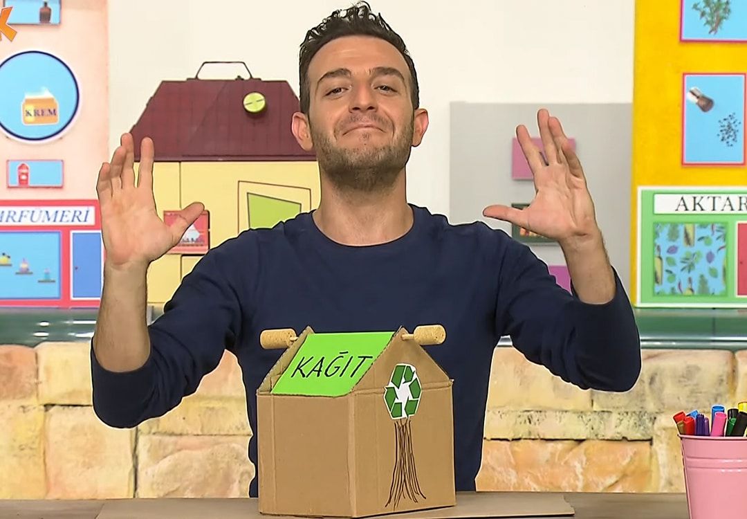 Geri dönüşüm kutusu video karesi: sunucu, üzerinde “KAĞIT” yazan ev biçimli karton kutunun arkasında