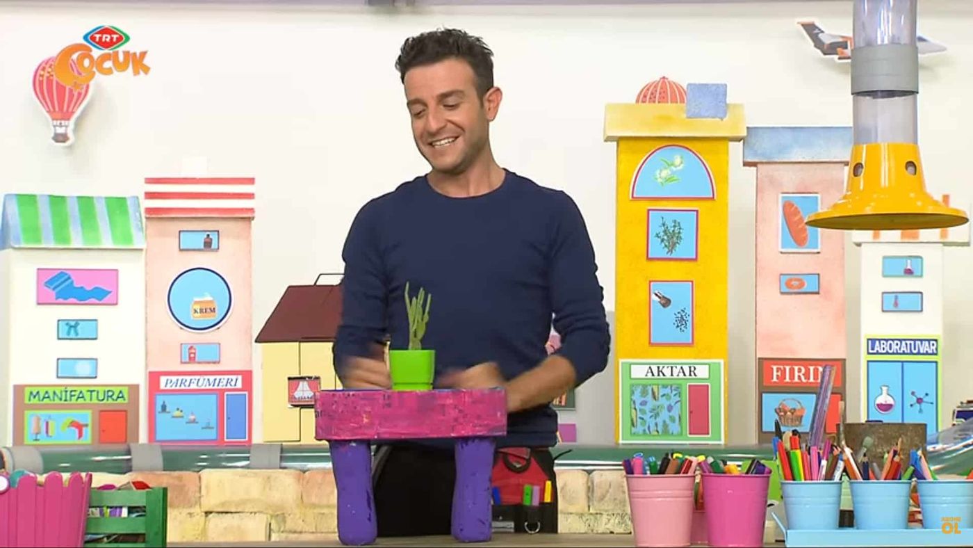 Stüdyoda sunucu, üstünde saksı duran mor ayaklı pembe sehpanın başında, video karesi