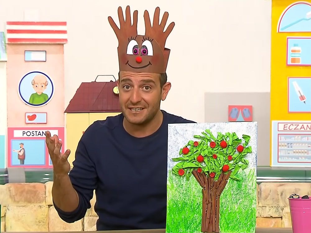 Geyik taç video karesi: sunucu başında el izi boynuzlu karton taç, yanında yeşil yapraklı ağaç tablosu