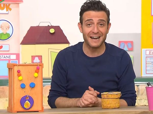 Televizyon programından kare: sunucu stüdyo masasında, yanında ahşap kutuya asılı seramik kolye