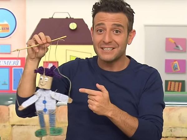 Karton Kukla: sunucu, mor yün saçlı ve ekose gömlekli ipli karton rulo kuklayı çubuğundan tutup gösteriyor
