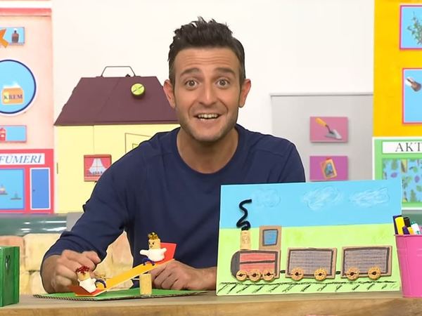 Televizyon programından kare: sunucu tahterevalli ve tren tablosunu masada gösteriyor