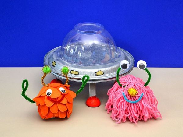 UFO ve sevimli uzaylılar: karton tabak ve şeffaf kubbeli gümüş UFO önünde turuncu ve pembe uzaylılar