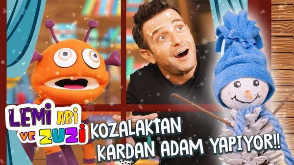 Kozalak kardan adam videosunun kapağı: sunucu, turuncu uzaylı kukla ve mavi bereli kozalak kardan adam