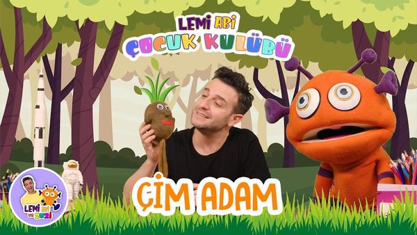 Çim adam video kapağı: orman arka planında sunucu elinde çim adam, yanında turuncu uzaylı kukla